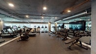 Located at 5750 Hollywood Blvd in the heart of Los Angeles, this fitness center offers a comprehensive workout facility designed to accommodate a wide range of training styles. The space features an industrial-modern aesthetic, with exposed concrete ceilings, visible ductwork, and recessed lighting that lend a sleek, urban character throughout.

The cardio zone is lined with treadmills, ellipticals, and cycling machines positioned near large windows that invite natural light and provide views of the surrounding greenery. A dedicated free weights area includes a fully stocked dumbbell rack with a wide range of weights, accompanied by adjustable benches and large wall-mounted mirrors for form monitoring. Flat-screen TVs are mounted on the walls, enhancing the workout experience.

The strength and functional training section features power racks, cable machines, barbells, and weight plates suited for serious resistance training. A built-in wooden storage unit houses medicine balls, foam rollers, yoga mats, and resistance tools, supporting stretching and functional fitness routines. Rubber flooring throughout the facility ensures safety and durability across all zones. The open, expansive layout allows for comfortable movement between stations, making this fitness center a thorough and well-appointed facility for all levels of fitness enthusiasts.
