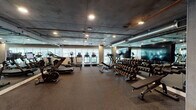 Located at 5750 Hollywood Blvd in the heart of Los Angeles, this fitness center offers a comprehensive workout facility designed to accommodate a wide range of training styles. The space features an industrial-modern aesthetic, with exposed concrete ceilings, visible ductwork, and recessed lighting that lend a sleek, urban character throughout.

The cardio zone is lined with treadmills, ellipticals, and cycling machines positioned near large windows that invite natural light and provide views of the surrounding greenery. A dedicated free weights area includes a fully stocked dumbbell rack with a wide range of weights, accompanied by adjustable benches and large wall-mounted mirrors for form monitoring. Flat-screen TVs are mounted on the walls, enhancing the workout experience.

The strength and functional training section features power racks, cable machines, barbells, and weight plates suited for serious resistance training. A built-in wooden storage unit houses medicine balls, foam rollers, yoga mats, and resistance tools, supporting stretching and functional fitness routines. Rubber flooring throughout the facility ensures safety and durability across all zones. The open, expansive layout allows for comfortable movement between stations, making this fitness center a thorough and well-appointed facility for all levels of fitness enthusiasts.