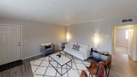 This single-floor residence offers a thoughtfully arranged layout that balances comfort and functionality across its key living spaces. The living room provides a welcoming, open atmosphere with wood-style flooring, neutral gray walls, and a textured ceiling. The room comfortably accommodates a seating arrangement centered around a geometric coffee table, with warm ambient lighting creating an inviting ambiance. Adjacent to the living area, the galley-style kitchen is efficiently designed with white cabinetry, light quartz-style countertops, a double stainless steel sink positioned beneath a window for natural light, and white appliances including an electric range and refrigerator. Wood-style flooring continues through the kitchen, adding visual continuity. The primary bedroom features plush carpet, soft gray walls, a window with blinds allowing natural light, and a striking drum chandelier with crystal accents as a centerpiece. The second bedroom mirrors a similarly cozy atmosphere, also carpeted, with its own elegant crystal chandelier and window, lending a refined character to the space. The bathroom is clean and functional, finished with wood-style flooring, a white vanity with sink, a large mirror, and a shower-tub combination with a curtain. Throughout, the neutral color palette and consistent finishes create a cohesive and modern aesthetic.