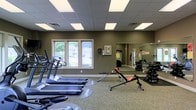 This fitness center offers a comprehensive workout environment thoughtfully designed to support a range of exercise routines. The space is finished with neutral-toned walls and a drop ceiling fitted with recessed panel lighting, ensuring consistent, even illumination throughout. A row of Vision Fitness treadmills and cardio machines lines one side of the room, positioned near a window that allows natural light to flow in while providing views of the surrounding greenery outside. An adjustable workout bench sits centrally, catering to a variety of strength training exercises. Along the opposite wall, a large floor-to-ceiling mirror expands the sense of space and allows users to monitor their form during workouts. A dedicated free weights area with a full dumbbell rack is reflected in the mirror, making the weight selection easily accessible. The flooring features a combination of carpet and exercise mat sections, providing traction and comfort underfoot. The overall ambiance is clean, functional, and organized, making it a practical and user-friendly environment for fitness enthusiasts of all levels.