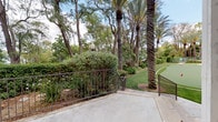 Located at 6021 Crape Myrtle Court in Los Angeles, CA, this meticulously maintained outdoor putting green offers a resort-style recreational experience set within a lush, private landscape. The putting surface features multiple holes marked with red flags, providing a functional and well-designed space for golf practice and leisure.

The green is framed by an impressive canopy of mature palm trees and a variety of broadleaf trees, creating a naturally shaded and visually striking environment. Dense, manicured hedgerows and ornamental plantings line the perimeter, enhancing the sense of privacy and seclusion. Decorative iron fencing and gated access points define the boundaries of the space while maintaining an open, airy feel.

Adjacent to the putting green is a charming patio area finished in warm-toned stonework, furnished with red accent seating and potted plants, offering a comfortable spot to relax before or after play. The nearby structure features a classic tile roof and stucco exterior, consistent with the Mediterranean-influenced architectural character of the property. A concrete walkway with exterior windows provides additional connectivity around the space.

Under the abundant Southern California sunshine, this outdoor area combines functional sports amenities with thoughtfully curated landscaping, making it an exceptional and inviting outdoor retreat.