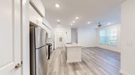 This single-floor unit offers a clean, modern design characterized by bright white walls, recessed lighting, and light wood-look flooring that runs consistently throughout the space. The open-concept layout seamlessly connects the kitchen and living area, creating a spacious, airy atmosphere enhanced by natural light from multiple windows fitted with white blinds.

The kitchen serves as a central hub of the unit, anchored by a large island with a granite countertop and an integrated sink. White shaker-style cabinetry lines the walls, complemented by a full suite of stainless steel appliances, including a refrigerator, gas range, and microwave. A built-in shelving nook and a dedicated desk area provide additional functional space within the kitchen zone.

The living area benefits from a ceiling fan and a window that draws in natural light. The bedrooms are well-proportioned, each featuring ceiling fans, large windows, and wood-look flooring. The primary bedroom includes access to a spacious walk-in closet outfitted with multi-level wire shelving systems for generous storage. A dedicated laundry area accommodates a full-size washer and dryer with overhead shelving. The overall aesthetic is clean, contemporary, and move-in ready.