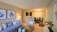 This single-floor residence showcases an open-concept design that seamlessly connects the living, dining, and kitchen areas into one cohesive, functional space. The living room is anchored by plush wall-to-wall carpeting in a warm neutral tone, complemented by soft beige walls that create an inviting and comfortable atmosphere. A sofa and a patterned accent chair provide ample seating, while a low-profile coffee table serves as a central gathering point. Coastal-inspired décor, including decorative vases and framed artwork, adds a relaxed, refined character to the space.

Adjacent to the living area, the dining space accommodates a round table surrounded by upholstered chairs, lit by a track light fixture overhead. Large double closet doors with white paneling provide generous storage directly in the dining area. A pass-through opening connects to the kitchen, which features dark cabinetry visible in the background, offering a sense of separation while maintaining the open flow of the layout. The overall ambiance is warm and welcoming, enhanced by layered lighting that includes both ambient and accent sources throughout the space.