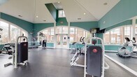 Located at 2000 Crossroads Manor Ct in Cary, NC, this single-floor fitness center is designed to provide a bright, motivating workout environment. The space is defined by its dramatic vaulted ceilings with a striking teal and white color scheme, lending a clean, energetic atmosphere throughout.

Large arched windows line multiple walls, allowing abundant natural light to fill the room while offering views of the surrounding community and greenery. The expansive rubber-floored workout area comfortably accommodates a well-rounded selection of equipment, including multiple treadmills, elliptical cross-trainers, a stationary bike, and several multi-station strength machines for full-body training. Wall-mounted flat-screen TVs are positioned throughout the space for entertainment during workouts.

Full-length mirrors along one wall visually expand the room and allow users to monitor their form during exercise. A ceiling fan provides additional air circulation, and recessed lighting supplements the natural light on overcast days. Double glass doors open directly to the exterior, enhancing ventilation and connectivity to the outdoors. The overall design prioritizes openness, functionality, and comfort, making this fitness center a well-equipped and inviting amenity for residents of the Crossroads Manor community.