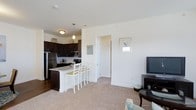 This two-floor residence offers a thoughtfully designed open-concept layout that seamlessly connects the kitchen, dining, and living areas. The kitchen is a standout feature, showcasing rich dark cabinetry, stainless steel appliances including a full-size refrigerator and built-in microwave, and a light-toned kitchen island with bar seating — ideal for casual dining or entertaining. Pendant lighting above the island adds a warm, inviting ambiance, complementing the hardwood-style flooring in the kitchen zone. The living area flows naturally from the kitchen, finished with plush neutral carpeting that adds comfort underfoot. Crisp white walls throughout the main level create a bright, airy atmosphere, enhanced by natural light from nearby windows. A framed wall piece and tasteful furnishings contribute to the space's clean, contemporary aesthetic. The open corridor connects additional rooms beyond the main living area, suggesting a practical and well-organized floor plan. Spread across two floors, this residence provides ample space for both everyday living and hosting, with a cohesive design language that balances modern functionality with a comfortable, livable feel.