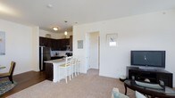 This two-floor residence offers a thoughtfully designed open-concept layout that seamlessly connects the kitchen, dining, and living areas. The kitchen is a standout feature, showcasing rich dark cabinetry, stainless steel appliances including a full-size refrigerator and built-in microwave, and a light-toned kitchen island with bar seating — ideal for casual dining or entertaining. Pendant lighting above the island adds a warm, inviting ambiance, complementing the hardwood-style flooring in the kitchen zone. The living area flows naturally from the kitchen, finished with plush neutral carpeting that adds comfort underfoot. Crisp white walls throughout the main level create a bright, airy atmosphere, enhanced by natural light from nearby windows. A framed wall piece and tasteful furnishings contribute to the space's clean, contemporary aesthetic. The open corridor connects additional rooms beyond the main living area, suggesting a practical and well-organized floor plan. Spread across two floors, this residence provides ample space for both everyday living and hosting, with a cohesive design language that balances modern functionality with a comfortable, livable feel.