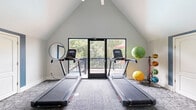 Located at 2814 De Mel Ave. in Louisville, KY, this dedicated fitness center is a thoughtfully designed exercise space that makes efficient use of its footprint. The room's most striking architectural feature is its soaring vaulted ceiling, which creates an airy, open atmosphere and adds a sense of spaciousness beyond the physical floor area.

The space is equipped with a comprehensive range of fitness equipment, including two treadmills, a recumbent stationary bike, a rowing machine, and a weight bench. A tiered dumbbell rack holds a full set of free weights, while an assortment of stability and medicine balls rounds out the functional training options. An exercise mat is mounted conveniently on the wall for easy access.

Patterned carpet tiles cover the floor throughout, providing both comfort and durability underfoot. A large glass-paned door opens to an outdoor deck, allowing natural light to flood the room and offering views of the surrounding greenery — a refreshing backdrop during workouts. A second glass door connects the fitness center to an adjacent common area, enhancing the sense of openness and flow.

An accent wall in deep teal anchors one side of the room and serves as a display area for posted fitness center rules, adding a pop of color to the otherwise neutral white interior. The space is clean, modern, and well-organized for a focused workout experience.