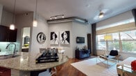 This single-floor urban residence showcases a contemporary loft-inspired design, highlighted by soaring ceilings with an exposed industrial-style duct that adds an artistic architectural element to the open-concept main living area. Rich hardwood flooring flows throughout, anchoring the space with warmth and continuity.

The living and dining areas are bathed in natural light from large windows dressed with elegant drapes, offering views of the surrounding streetscape. Bold black-and-white artwork adorns the walls, complementing the modern aesthetic, while track lighting and a ceiling fan provide functional yet stylish illumination.

The gourmet kitchen is a standout feature, boasting dark espresso cabinetry that reaches toward the ceiling, stone-look tile backsplash, stainless steel appliances including a gas range, and a sweeping granite-topped island with bar seating — perfect for casual dining or entertaining. Pendant lighting above the island adds a refined finishing touch.

The bedroom is a serene retreat, featuring plush carpeting, large windows with decorative curtains, and tasteful artwork displayed above a tufted upholstered bed frame. The bathroom is finished with large-format dark tile surrounding a full soaking tub, a dark-framed vanity mirror, and clean contemporary fixtures, delivering a spa-like atmosphere.