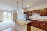 Located at 1300 Post Rd in Fitchburg, Wisconsin, this one-bedroom, one-bathroom unit offers a well-organized layout across a single primary floor. The open-concept kitchen and living area serves as the heart of the home, where rich, warm-toned wood cabinetry pairs with a granite-topped center island illuminated by elegant pendant lighting. Stainless steel appliances — including a refrigerator, dishwasher, and range — complete the kitchen, which also features a double sink and ample upper and lower cabinet storage. Light hardwood-style flooring runs through the kitchen and dining area, while plush carpeting extends into the living room and bedroom. A sliding glass door in the living area invites natural light and provides access to a private patio with outdoor views. The spacious bedroom offers a comfortable, carpeted retreat with a well-sized window for additional natural light. The bathroom is well-appointed with a wide vanity, generous wood cabinetry, a large mirror with lighted fixtures above, and durable flooring. Two closets provide practical storage throughout the unit, and a dedicated laundry area adds everyday convenience. The overall ambiance is clean, warm, and functional.