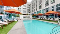 Located at 1509 Broadway in San Diego's vibrant Makers Quarter neighborhood, The Wyatt presents a thoughtfully designed two-level outdoor amenity space that embodies Southern California resort living. The centerpiece is a generously proportioned swimming pool with crystal-clear blue water, complemented by a separate spa, both finished in clean concrete surrounds that reflect the building's modern aesthetic.

Flanking the pool are expansive turf lounging areas outfitted with rows of wooden-framed chaise lounges, each accented with plush cushions and pillows. Bold orange cantilever umbrellas provide ample shade throughout the deck, adding a vibrant pop of color against the building's crisp white and glass facade. The surrounding multi-story residential building wraps the courtyard in a U-shaped configuration, creating a sense of enclosure while offering open sky views above.

A lower-level courtyard area features lush tropical palm plantings, glass railings, and comfortable outdoor seating arrangements, providing an additional social gathering space with a more intimate, garden-like atmosphere. Floor-to-ceiling glazing along the building's ground-floor facade connects the interior to the outdoor amenity areas seamlessly. The overall design balances an urban architectural language with resort-inspired comfort, offering residents a distinctive outdoor retreat in the heart of downtown San Diego.