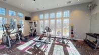 This fitness center is designed to support a comprehensive workout experience within a bright, open, and inviting environment. The space is characterized by soaring ceilings and a generous wall of large, grid-paned windows that span nearly the full height of the room, allowing abundant natural light to fill the interior and offering views of the surrounding outdoor landscape.

The flooring features a bold, graphic-patterned carpet in shades of brown, burgundy, pink, and gray, adding a dynamic visual element to the space. Recessed lighting throughout the ceiling provides consistent illumination, complementing the natural light.

Cardio equipment is well-represented, including treadmills, elliptical trainers, and stationary bikes, positioned to take full advantage of the outdoor views. Strength training equipment includes a cable machine, a multi-functional weight station, a dumbbell rack with a full set of free weights, adjustable benches, and a medicine ball rack stocked with a colorful assortment of weighted balls. Wall-mounted televisions are positioned throughout for entertainment during workouts.

Large mirrors on one wall enhance the sense of space and provide functional support for strength training. A wall-mounted fan ensures proper air circulation. The overall ambiance is clean, modern, and energizing, making this fitness center a well-rounded facility suitable for a wide range of exercise routines.