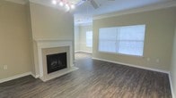 This single-floor residence offers a thoughtfully designed living space that balances comfort and classic style. The main living area immediately captures attention with its tile-surround fireplace, serving as a natural focal point and adding warmth to the room. Rich, hardwood-style flooring extends throughout, providing a durable yet elegant foundation that ties the space together seamlessly. Crown molding runs along the upper walls, lending a refined architectural detail that elevates the overall aesthetic. The room is bathed in natural light, courtesy of large windows dressed with white horizontal blinds, creating a bright and airy atmosphere. A ceiling fan with a built-in light fixture ensures comfort year-round, while a track lighting fixture adds additional illumination. The neutral, warm-toned walls provide a versatile backdrop, making the space easy to personalize. The open layout allows for flexible furniture arrangements, accommodating both relaxed everyday living and entertaining with ease. Crisp white baseboards and clean lines throughout reinforce the space's well-maintained and move-in-ready appeal, making it an inviting and functional home.