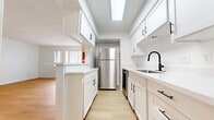 This two-story condo at 3411 Cummins St spans 1,000 square feet across two levels. The main floor (650 sq ft) features a bright living room with light hardwood-style flooring, built-in shelving, and a carpeted staircase leading upstairs. The kitchen is outfitted with white shaker cabinets, black hardware, light countertops, and stainless steel appliances. Upstairs (350 sq ft), both bedrooms feature ceiling fans and carpet. The 200 sq ft primary bedroom includes mirrored sliding closet doors and built-in shelving, while a secondary bedroom offers balcony access with urban views. The bathrooms feature white cabinetry with light countertops. A shared outdoor balcony provides open city views.