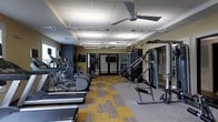This fitness center offers a comprehensive workout environment housed within a single-floor layout designed for both cardio and strength training. The space is outfitted with a lineup of cardio machines, including treadmills and elliptical trainers, arranged in a row along one side to allow for comfortable, unobstructed use. On the opposite side, a multi-station cable and weight machine provides options for a full range of resistance exercises, complemented by additional strength equipment. The flooring features a patterned carpet tile in a combination of yellow and gray tones, adding a modern, energetic aesthetic to the space. Overhead, a drop ceiling with recessed and linear lighting ensures the room is well-lit throughout, while a ceiling fan aids in air circulation during intense workouts. The walls are painted in a warm neutral tone, and mirrored panels near the back of the room enhance the sense of openness and allow users to monitor their form. A set of glass-paneled doors at the rear provides access to an adjacent area, contributing to the overall functionality and flow of the facility. The space is thoughtfully arranged to maximize usability while maintaining a clean and organized environment.