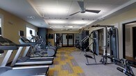 This fitness center offers a comprehensive workout environment housed within a single-floor layout designed for both cardio and strength training. The space is outfitted with a lineup of cardio machines, including treadmills and elliptical trainers, arranged in a row along one side to allow for comfortable, unobstructed use. On the opposite side, a multi-station cable and weight machine provides options for a full range of resistance exercises, complemented by additional strength equipment. The flooring features a patterned carpet tile in a combination of yellow and gray tones, adding a modern, energetic aesthetic to the space. Overhead, a drop ceiling with recessed and linear lighting ensures the room is well-lit throughout, while a ceiling fan aids in air circulation during intense workouts. The walls are painted in a warm neutral tone, and mirrored panels near the back of the room enhance the sense of openness and allow users to monitor their form. A set of glass-paneled doors at the rear provides access to an adjacent area, contributing to the overall functionality and flow of the facility. The space is thoughtfully arranged to maximize usability while maintaining a clean and organized environment.