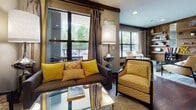 Located at 4000 City Walk Way in Charlottesville, Virginia, this single-floor communal space combines contemporary design with warm, inviting elements to create a multifunctional gathering environment. Rich, dark hardwood flooring flows continuously throughout, anchoring the space with a sense of warmth and cohesion. Exposed wood beam accents along the ceiling and walls add a rustic touch that complements the modern furnishings.

The lounge area features plush upholstered sofas and chairs in bold golden yellow tones paired with chevron-patterned textiles, arranged around a mirrored coffee table beneath large windows with floor-length drapes that allow generous natural light. A built-in linear fireplace set against a textured stone accent wall creates a focal point in a quieter seating nook, accompanied by pendant lighting and a cushioned banquette.

A well-appointed kitchenette includes dark cabinetry, stainless steel appliances, a wall-mounted flat-screen TV framed in yellow, and ample counter space. An integrated workstation with floating shelves and a dedicated desk chair provides a functional workspace. Decorative open shelving displays curated accessories throughout, and glass double doors offer a welcoming entry point, completing this thoughtfully designed communal environment.
