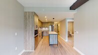 This single-floor unit showcases a cohesive modern design, highlighted by light wood-tone laminate flooring that flows seamlessly throughout the space. The kitchen serves as the centerpiece, featuring wood-grain cabinetry extending floor to ceiling, stainless steel appliances including a range and refrigerator, and a white subway tile backsplash. A freestanding island with a light countertop provides additional prep and seating space, while recessed and pendant lighting illuminate the area effectively. Black hardware accents on cabinetry and fixtures add a contemporary contrast throughout.

The unit includes two bathrooms, each finished with large-format tile and matte black fixtures. One bathroom features a glass-enclosed walk-in shower with mosaic tile flooring and grab bars, while the second offers a full tub-shower combination with tiled surround, an undermount sink, and an illuminated LED mirror set above wood-grain vanity cabinetry.

The bedroom is a well-proportioned, open room bathed in natural light through a large picture window offering urban views. Neutral warm-gray walls complement the light flooring, creating a calm and versatile environment. The overall design of this unit balances functionality and contemporary aesthetics with clean lines and quality finishes throughout.
