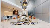 Located at 2410 Canopy Breeze Circle in Orlando, Florida, this single-floor community clubhouse and leasing center is a showcase of contemporary interior design and architectural sophistication. The grand entrance lobby commands immediate attention with its soaring double-height ceiling, geometric coffered panels, and dramatic sculptural pendant light fixtures in black and gold that draw the eye upward. Large-format white marble-look porcelain tile flows throughout, anchored by vibrant, abstract area rugs in bold primary colors that add artistic energy to the space.

The reception area features a sleek, curved white desk adorned with cascading gold globe pendant lights, framed by curated wall art and decorative metallic accents. Adjacent lounge areas offer comfortable seating arrangements in neutral tones accented with rich orange and navy throw pillows. Floor-to-ceiling glass walls at the rear of the lobby frame breathtaking views of a resort-style pool surrounded by palm trees, seamlessly connecting the interior to the outdoor amenity space.

The leasing suite transitions into a richly appointed lounge with a slate-blue wood-panel ceiling, built-in display shelving, an oxidized botanical wall feature, and a co-working station. A dedicated business center and private consultation rooms extend down a bright, marble-tiled corridor, completing this thoughtfully designed, amenity-rich facility.