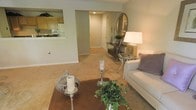 This single-story residence at 9723-A offers a thoughtfully designed layout with an open and airy feel. The living area is carpeted and comfortably sized, providing a relaxed setting accented by neutral wall tones and clean white baseboards. A decorative coffee table with candle accents and greenery anchors the seating area, which flows naturally toward a defined entryway. The entry features a console table topped with a round mirror and a potted plant, creating a stylish focal point upon arrival.

Adjacent to the living space, a kitchen pass-through with warm wood cabinetry and white appliances offers convenient sight lines between the kitchen and main living area, making the layout feel connected and functional. The recessed lighting and soft, neutral palette throughout contribute to a calm, welcoming ambiance. The open corridor leading toward the entryway door adds depth to the space and reinforces the single-floor layout's easy flow. Overall, the unit presents a clean, well-maintained interior with classic finishes suitable for comfortable everyday living.