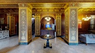 This magnificent lobby is a showcase of classical architectural grandeur, defined by its richly detailed ornamental murals that line the walls in vibrant golds, greens, and earthy tones. Elaborate painted panels feature symmetrical motifs of urns, floral arrangements, and portrait medallions framed within gilded borders, reflecting a refined artistic tradition. The coffered ceiling continues this opulence, adorned with intricate geometric and floral patterns in warm gold tones that draw the eye upward and lend the space an impressive sense of height and formality. Polished terrazzo flooring adds a refined, durable foundation that complements the overall classical aesthetic. At the center of the space, a dark wood pedestal table serves as an elegant focal point, accented by a bouquet of fresh yellow flowers that adds a welcoming touch of color. A graceful arched passageway leads to a wood-paneled reception desk, fitted with a monitor and framed by a chandelier overhead, providing a functional yet stylish service area. To one side, a staircase extends the space vertically, hinting at additional areas beyond. The overall ambiance is one of historic sophistication and institutional prestige, evoking the character of a landmark building steeped in architectural heritage.