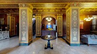 This magnificent lobby is a showcase of classical architectural grandeur, defined by its richly detailed ornamental murals that line the walls in vibrant golds, greens, and earthy tones. Elaborate painted panels feature symmetrical motifs of urns, floral arrangements, and portrait medallions framed within gilded borders, reflecting a refined artistic tradition. The coffered ceiling continues this opulence, adorned with intricate geometric and floral patterns in warm gold tones that draw the eye upward and lend the space an impressive sense of height and formality. Polished terrazzo flooring adds a refined, durable foundation that complements the overall classical aesthetic. At the center of the space, a dark wood pedestal table serves as an elegant focal point, accented by a bouquet of fresh yellow flowers that adds a welcoming touch of color. A graceful arched passageway leads to a wood-paneled reception desk, fitted with a monitor and framed by a chandelier overhead, providing a functional yet stylish service area. To one side, a staircase extends the space vertically, hinting at additional areas beyond. The overall ambiance is one of historic sophistication and institutional prestige, evoking the character of a landmark building steeped in architectural heritage.