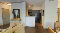This single-floor residence offers a thoughtfully designed open-concept layout that connects the living area and kitchen in a cohesive, functional arrangement. Rich hardwood-style flooring runs throughout the space, lending warmth and continuity from room to room. The living area provides a comfortable setting, highlighted by clean white walls that enhance the sense of openness and brightness. A decorative console table with a framed mirror adds a stylish accent near the entryway transition.

The kitchen is positioned at the heart of the home, separated from the living space by a half-wall peninsula that doubles as a breakfast bar. Dark gray cabinetry lines the kitchen walls, offering ample storage and a modern aesthetic, while a tile backsplash adds texture and visual interest. Black appliances, including a full-size refrigerator and microwave, complement the cabinet tones for a cohesive, contemporary look. Track lighting above the kitchen illuminates the workspace effectively.

The corridor leading further into the residence hints at additional private spaces beyond, with crisp white walls and consistent flooring maintaining the clean, modern aesthetic throughout. Overall, the space balances contemporary design with practical functionality.