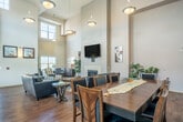 Located at 1061 Acker Ln in Verona, Wisconsin, this single-floor community space offers a welcoming and well-appointed environment that seamlessly combines a living room and dining area. The most striking architectural feature is the dramatic two-story ceiling height, which lends an expansive, airy quality throughout the entire space. Warm hardwood flooring runs continuously underfoot, complementing the neutral, earthy tones of the walls and creating a cohesive, inviting atmosphere.

The living area is anchored by a tile-surround gas fireplace with a wall-mounted flat-screen television above it, flanked by navy blue leather sofas arranged around a round coffee table — ideal for relaxed gathering. Pendant lighting hangs from the soaring ceiling, adding warmth and visual interest. Multiple large windows positioned at both standard and upper clerestory levels flood the room with natural light and offer views of the surrounding Verona landscape.

The dining area features a substantial high-top table with upholstered teal chairs, accompanied by additional bar-height seating. A large window frames an outdoor view, and wall sconces provide soft ambient lighting. Lush potted plants are thoughtfully placed throughout, adding a natural, refreshing touch. A glass door provides additional access to the exterior, further enhancing the connection between indoors and outdoors.