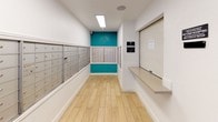 The mail area at Bella Vista at Hilltop is a compact, single-floor space designed to efficiently serve residents' postal needs. The layout is straightforward and functional, with light wood-style plank flooring running the length of the corridor, contributing a clean and modern aesthetic throughout.

The most prominent feature is the extensive bank of individual resident mailboxes lining the walls on multiple sides. These standard metal cluster mailbox units are organized in rows, providing dedicated mail storage for a large number of residents. A vivid teal accent wall anchors the far end of the space, adding a distinctive pop of color against the otherwise white walls and trim.

A built-in counter with a roll-up shutter is positioned along one wall, functioning as a package or parcel service window for residents. Overhead, recessed and flush-mounted lighting fixtures ensure the space is well-lit. A glass-paned entry door opens directly to an outdoor walkway and landscaped grounds, allowing natural light to filter in and offering a view of the surrounding community. A small waste receptacle is conveniently placed near the entrance. Security is supported by a visible camera mounted near the ceiling.