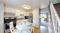This two-floor residence offers a thoughtfully designed layout with a clean, neutral aesthetic that flows seamlessly from room to room. The kitchen is a functional highlight, featuring white shaker-style cabinetry, dark granite-look countertops, stainless steel appliances including a gas range and refrigerator, and warm hardwood-style flooring. A compact dining area adjacent to the kitchen accommodates a round white table, making it an ideal space for casual meals. The kitchen opens toward a comfortable living room finished with plush carpeting and large sliding glass doors that invite an abundance of natural light and offer pleasant views of the surrounding greenery. The living room provides a welcoming atmosphere with ample space for seating and relaxation. The bedrooms are carpeted and well-proportioned, offering a serene retreat with neutral wall tones. One bedroom features access to a dedicated closet space, providing practical storage. The bathroom is clean and functional, equipped with a full-size soaking tub and shower combination, white tile flooring, and simple, modern fixtures. Throughout the residence, the consistent use of light gray and white tones, paired with warm wood accents, creates a cohesive and inviting environment suited for comfortable everyday living.