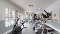 The fitness center at Cambridge Apartments is a thoughtfully designed amenity space that provides residents with a comprehensive workout environment. The room features a neutral color palette of grey and white, accented by crown molding and a textured ceiling, lending a polished, residential feel to the space.

Natural light is a defining characteristic of this fitness center, with multiple French doors and windows lining the perimeter walls. These glazed openings offer views of the outdoor surroundings, creating an open, airy atmosphere that enhances the workout experience. Recessed lighting and wall-mounted sconces supplement natural light, ensuring the space remains well-lit throughout the day.

The facility is outfitted with a diverse range of equipment to support both cardiovascular and strength training. Cardio machines include ellipticals, treadmills, and a recumbent stationary bike, arranged along one side of the room. A multi-station cable weight machine serves as the centerpiece for strength training, complemented by a rack of free weights and kettlebells. A large wall mirror expands the sense of space and allows users to monitor their form. A wall-mounted flat-screen television adds convenience for entertainment during workouts. Dark-toned flooring with protective rubber mats completes the functional, well-appointed space.