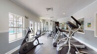 The fitness center at Cambridge Apartments is a thoughtfully designed amenity space that provides residents with a comprehensive workout environment. The room features a neutral color palette of grey and white, accented by crown molding and a textured ceiling, lending a polished, residential feel to the space.

Natural light is a defining characteristic of this fitness center, with multiple French doors and windows lining the perimeter walls. These glazed openings offer views of the outdoor surroundings, creating an open, airy atmosphere that enhances the workout experience. Recessed lighting and wall-mounted sconces supplement natural light, ensuring the space remains well-lit throughout the day.

The facility is outfitted with a diverse range of equipment to support both cardiovascular and strength training. Cardio machines include ellipticals, treadmills, and a recumbent stationary bike, arranged along one side of the room. A multi-station cable weight machine serves as the centerpiece for strength training, complemented by a rack of free weights and kettlebells. A large wall mirror expands the sense of space and allows users to monitor their form. A wall-mounted flat-screen television adds convenience for entertainment during workouts. Dark-toned flooring with protective rubber mats completes the functional, well-appointed space.