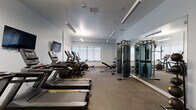 This fitness center at 401 A St in Wilmington, Delaware, is a thoughtfully designed exercise facility that packs a comprehensive range of workout equipment into a compact, efficiently organized single-room space. The room features light gray walls, a clean white ceiling with recessed linear pendant lighting, and durable carpet-tiled flooring suited for high-traffic fitness use.

Along one side of the room, a row of treadmills lines the wall beneath a wall-mounted flat-screen television, providing an engaging cardio experience. Adjacent to these are multiple elliptical trainers and a stationary bike, offering varied low-impact cardio options. A Matrix-brand adjustable weight bench provides a dedicated area for strength exercises. On the opposite side, a multi-functional cable machine and strength training tower are positioned in front of a large floor-to-ceiling mirror, allowing users to monitor their form during workouts.

Additional accessories include a selection of stability/medicine balls neatly stored on a rack near the entrance, alongside a small shelving unit with fitness accessories. Natural light filters in through several roller-shade windows along the rear wall, creating a bright and energizing atmosphere. The space is accessed through double glass-panel doors, lending a polished, professional feel to this well-appointed fitness facility.