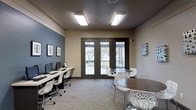 This single-floor space is designed as a functional and modern shared workspace, blending productivity-focused amenities with a welcoming atmosphere. The room features a dual-toned color scheme, with a deep slate-blue accent wall complementing neutral beige tones, creating a professional yet approachable environment. Recessed rectangular flush-mount lighting panels illuminate the space evenly from a dark ceiling, contributing to a clean, contemporary aesthetic.

Along one wall, a linear workstation desk accommodates multiple computer monitors and keyboards, paired with sleek white task chairs — ideal for focused individual work. The opposite side of the room features two round dark-wood tables surrounded by ornate white lattice-design chairs, offering comfortable spots for casual meetings or group collaboration.

A set of dark-framed double glass doors at the center of the far wall floods the room with natural light and provides a view of an outdoor area with mature trees, adding a refreshing visual connection to the exterior. Framed artwork adorns the walls, adding subtle character to the space. Neutral carpeted flooring ties the room together, and the overall layout balances utility with a polished, community-oriented design.