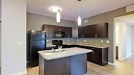 This single-floor space showcases a thoughtfully designed interior with a strong emphasis on modern aesthetics. The kitchen serves as a central highlight, featuring rich dark espresso cabinetry that extends from floor to ceiling, providing generous storage throughout. A central island anchors the space, equipped with a double stainless steel sink and offering additional workspace and casual seating opportunities. The countertops carry a neutral stone-toned finish that complements the dark cabinetry, while the light hardwood-style flooring adds warmth and contrast to the overall palette. Black appliances, including a full-size refrigerator, range, and built-in microwave, maintain a cohesive, contemporary look. Overhead, a combination of pendant lights and a flush-mount ceiling fixture provides layered illumination, with two sleek drop pendants drawing focus to the island and creating an inviting ambiance. The kitchen layout follows a practical U-shape configuration, maximizing counter space and workflow efficiency. Neutral gray walls tie the design elements together, creating a clean, modern atmosphere throughout the space. Ventilation is provided through ceiling-integrated HVAC vents, ensuring comfort year-round.