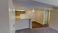 This single-floor residence offers a practical and comfortable open-concept layout that connects the kitchen and living areas in a cohesive, functional design. The kitchen is defined by warm wood-toned upper cabinetry and a built-in sink, all illuminated by a recessed fluorescent light fixture above. A peninsula-style breakfast bar serves as a natural divider between the kitchen and the adjoining living space, providing both a casual dining surface and an open sightline throughout the common areas. The transition from the carpeted entryway zone to the rich, dark hardwood flooring of the living room creates a clear visual distinction between spaces while maintaining an overall sense of flow. The living area is generously proportioned, featuring neutral warm-toned walls that lend a welcoming ambiance. A ceiling fan with a built-in light fixture is centrally positioned, offering both ventilation and ambient lighting. White trim and baseboards throughout provide clean contrast against the warm wall tones. A doorway visible at the far end of the living area leads to additional rooms, suggesting a well-connected interior layout. The space is well-maintained, straightforward in its design, and offers a comfortable, move-in ready environment.