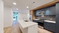Located at 1150 Spring St NW in Atlanta's vibrant Midtown neighborhood, this contemporary single-floor unit showcases a thoughtfully designed open layout with clean architectural lines and high-quality finishes throughout. Light wood-tone flooring flows seamlessly across the main living areas, complementing the bright white walls and recessed lighting that give the space an airy, modern feel.

The kitchen is a standout feature, centered around a generous island with a white quartz countertop and an undermount sink with a professional-style faucet. Sleek navy-blue cabinetry with integrated hardware lines the walls, accented by warm wood upper panels and a textured gray tile backsplash. Stainless steel appliances, including a range, microwave, and French door refrigerator, complete the upscale culinary space.

The open living area extends naturally from the kitchen, offering a versatile and well-proportioned space bathed in natural light. The bedroom features plush carpet flooring and floor-to-ceiling sliding glass windows that frame striking views of the surrounding Atlanta cityscape, flooding the room with natural light.

The bathroom is finished with dark navy cabinetry, a white quartz vanity, backlit mirrors, and wood-look luxury vinyl flooring. A soaking tub with a bold dark tile surround and a curved chrome shower rod adds a refined, contemporary touch.