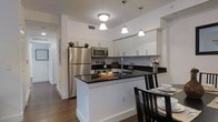 This single-floor residence offers a thoughtfully designed living space with a clean, modern aesthetic. The kitchen serves as a standout feature, showcasing white shaker-style cabinetry that extends to the ceiling, providing ample storage throughout. Dark granite countertops create a striking contrast against the bright white surfaces, lending the space a polished, contemporary feel. A central island with matching granite offers additional prep space and doubles as a casual dining or entertaining area, illuminated by a trio of pendant lights that hang at varying heights above. Stainless steel appliances, including a full-size refrigerator, built-in microwave, and range, complete the kitchen's modern profile. Warm hardwood flooring runs throughout, adding texture and a sense of continuity to the space. The layout flows naturally toward a dedicated dining area, where a dark wood table and chairs complement the kitchen's color palette. Recessed lighting and pendant fixtures ensure the space is well-lit, while the bright white walls and clean lines contribute to an open, airy ambiance. A hallway visible from the kitchen suggests additional rooms beyond, completing this well-organized and stylish residence.