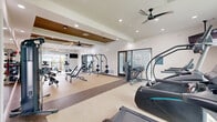 Located in Port Saint Lucie, Florida, this modern fitness center offers a comprehensive, multi-zone workout facility spread across a single floor. The space is thoughtfully organized into distinct areas designed to support a full range of exercise modalities.

The main strength and free weights area features an impressive array of equipment, including cable machines, barbells, weight racks, adjustable benches, dumbbells, and medicine balls. This zone is anchored by a striking tray ceiling with warm bronze accents, ceiling fans, and geometric pendant lighting, lending an upscale aesthetic to the workout environment. Full-length mirrors line the walls, enhancing the sense of space.

A dedicated cardio zone houses treadmills, ellipticals, stair climbers, and a WaterRower, with large windows that invite abundant natural light and outdoor views of the surrounding community. An open-air section features a retractable overhead garage door that connects the cardio area to a balcony, providing fresh-air workout options.

A separate functional fitness room offers wood-look flooring, stability balls, cycling bikes, and a large mirror wall — ideal for stretching, cycling, or guided workouts. Adjacent to it is a bright, comfortable lounge area with soft seating, a marble-top table, and a glass wall overlooking the fitness floor, providing a welcoming space to rest and recover.