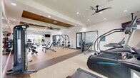 Located in Port Saint Lucie, Florida, this modern fitness center offers a comprehensive, multi-zone workout facility spread across a single floor. The space is thoughtfully organized into distinct areas designed to support a full range of exercise modalities.

The main strength and free weights area features an impressive array of equipment, including cable machines, barbells, weight racks, adjustable benches, dumbbells, and medicine balls. This zone is anchored by a striking tray ceiling with warm bronze accents, ceiling fans, and geometric pendant lighting, lending an upscale aesthetic to the workout environment. Full-length mirrors line the walls, enhancing the sense of space.

A dedicated cardio zone houses treadmills, ellipticals, stair climbers, and a WaterRower, with large windows that invite abundant natural light and outdoor views of the surrounding community. An open-air section features a retractable overhead garage door that connects the cardio area to a balcony, providing fresh-air workout options.

A separate functional fitness room offers wood-look flooring, stability balls, cycling bikes, and a large mirror wall — ideal for stretching, cycling, or guided workouts. Adjacent to it is a bright, comfortable lounge area with soft seating, a marble-top table, and a glass wall overlooking the fitness floor, providing a welcoming space to rest and recover.