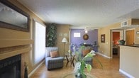 This single-floor residence offers a welcoming and well-organized layout that combines comfort with character. The open-concept living and dining area serves as the heart of the home, seamlessly connecting everyday living spaces. Warm golden-beige walls set a cozy tone throughout, while a bold purple accent wall in the dining area creates a striking visual focal point that adds personality and depth to the space.

The living area is furnished with plush, striped armchairs and accented with a glass side table, creating a relaxed and inviting atmosphere. A tall floor lamp and lush indoor greenery contribute to the warm, homey feel. Large vertical window treatments allow natural light to filter in, brightening the space throughout the day.

The dining area features a ceiling fan with a light fixture, providing both comfort and illumination. A decorative mirror adorns the purple accent wall, complementing the dining set beneath it. Hardwood-style flooring runs throughout, adding warmth and visual continuity across both areas.

A textured popcorn ceiling adds a classic touch overhead, tying together the overall aesthetic of this cozy, characterful residence. The thoughtful use of color, natural elements, and open sightlines makes this a practical yet visually engaging space.