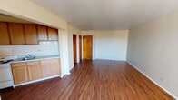 Located at 7042 S South Shore Dr in Chicago's South Shore neighborhood, this single-floor unit presents a straightforward, functional layout with consistent wood-style flooring throughout. The open living area is generously proportioned, featuring neutral walls and a large window that allows natural light to fill the space while providing outdoor views. A baseboard radiator provides heating beneath the window.

The kitchen is positioned in an alcove adjacent to the main living area, offering an efficient and accessible design. It features medium-toned wood upper cabinets and lighter lower cabinetry, a tile backsplash, a white range, sink, and a full-size refrigerator. The semi-open configuration keeps the kitchen connected to the living space.

A hallway connects the living area to additional spaces, including a closet with a built-in shelf and hanging rod, providing practical storage. The bathroom includes tile flooring in a classic mosaic pattern, a vanity with a mirror, and white tiled walls with wainscoting detail extending into the adjacent hallway. White trim and clean lines contribute to a simple, well-maintained aesthetic throughout the unit.