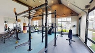 Located at 1062 W Oakland Ave in Johnson City, Tennessee, this fitness center offers a thoughtfully designed exercise space that is both functional and visually impressive. The single-floor layout maximizes its generous footprint, providing ample room for a diverse range of workout activities.

Architecturally, the space is defined by soaring vaulted ceilings with warm wood paneling and exposed beams that lend a modern yet inviting aesthetic. Large windows along one wall and a full-width glass garage door flood the interior with natural light, offering views of the outdoor green space and pool area beyond. The open garage door design allows for seamless indoor-outdoor connectivity.

The exercise room is comprehensively outfitted with a wide array of equipment. Cardio options include multiple treadmills, elliptical machines, a stair climber, and Concept2 rowing ergometers. A dedicated free weights section features a full rack of dumbbells accompanied by adjustable benches. Strength training is further supported by a multi-functional power rack and a cable machine system. A hanging heavy punching bag adds a combat fitness element to the space. Full-length mirrors along one wall enhance the sense of openness while allowing users to monitor their form. The flooring transitions between teal carpet and darker rubberized surfaces, delineating functional zones throughout the room.