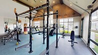 Located at 1062 W Oakland Ave in Johnson City, Tennessee, this fitness center offers a thoughtfully designed exercise space that is both functional and visually impressive. The single-floor layout maximizes its generous footprint, providing ample room for a diverse range of workout activities.

Architecturally, the space is defined by soaring vaulted ceilings with warm wood paneling and exposed beams that lend a modern yet inviting aesthetic. Large windows along one wall and a full-width glass garage door flood the interior with natural light, offering views of the outdoor green space and pool area beyond. The open garage door design allows for seamless indoor-outdoor connectivity.

The exercise room is comprehensively outfitted with a wide array of equipment. Cardio options include multiple treadmills, elliptical machines, a stair climber, and Concept2 rowing ergometers. A dedicated free weights section features a full rack of dumbbells accompanied by adjustable benches. Strength training is further supported by a multi-functional power rack and a cable machine system. A hanging heavy punching bag adds a combat fitness element to the space. Full-length mirrors along one wall enhance the sense of openness while allowing users to monitor their form. The flooring transitions between teal carpet and darker rubberized surfaces, delineating functional zones throughout the room.