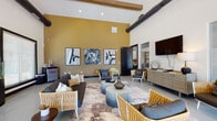 This single-floor communal space combines a stylish lounge area with an open game room, unified by dramatic soaring ceilings accented with exposed wooden beams and industrial ductwork. The lounge area is anchored by a bold golden-yellow accent wall that serves as a gallery backdrop for a trio of large abstract artworks in black frames. Polished concrete floors flow throughout, lending an industrial-modern character to the space. Seating is arranged around cylindrical concrete coffee tables and includes woven accent chairs in warm gold and white tones paired with deep charcoal sofas, creating an inviting yet contemporary aesthetic. A wall-mounted television and a neutral-toned credenza add functional elements, while a compact beverage refrigerator provides added convenience.

The game room section opens dramatically via large glass garage-style doors, flooding the area with natural light and offering views of the surrounding outdoors. A centerpiece pool table with a white frame and dark felt surface commands the space, accompanied by ample room for play. A built-in aquarium adds a distinctive focal point along one wall, while additional seating and a wall-mounted television complete the entertainment-oriented layout. Together, the zones create a cohesive, energetic environment well-suited for socializing and recreation.