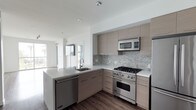 This single-floor space showcases a thoughtfully designed, open-concept layout anchored by a modern kitchen. The kitchen is outfitted with flat-front cabinetry in a warm taupe finish, complemented by brushed metal hardware that adds a refined, contemporary touch. A mosaic tile backsplash in neutral grey tones runs along the cooking wall, providing subtle texture and visual interest.

Premium stainless steel appliances are a standout feature, including a professional-grade gas range, built-in microwave, dishwasher, and refrigerator — offering both style and functionality. The kitchen island provides additional workspace and accommodates an undermount sink, making it both a practical and social hub of the space.

Dark hardwood flooring grounds the kitchen and contrasts beautifully with the bright white countertops and light-toned cabinetry. Recessed ceiling lighting ensures the kitchen is well-illuminated throughout. The open sightlines from the kitchen into the adjacent living area, enhanced by large windows that invite natural light, create a bright and cohesive atmosphere throughout the space.