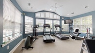 This dedicated fitness center is thoughtfully designed to support a full range of exercise routines within a bright, welcoming environment. The space features striking vaulted ceilings that lend a sense of openness and volume, complemented by a bold steel-blue color palette on the walls and crisp white trim throughout.

The main workout area is generously sized and bathed in natural light, courtesy of a wide bank of large windows dressed with white plantation shutters. These windows frame pleasant outdoor views of surrounding greenery, creating an energizing atmosphere for exercise. A ceiling fan overhead ensures comfortable air circulation, while recessed lighting provides additional illumination.

Cardio equipment is prominently featured, with multiple treadmills, an elliptical trainer, and a stationary bike arranged to take full advantage of the natural light and views. A wall-mounted flat-screen television adds entertainment options for cardio sessions. Strength training needs are well served by a multi-station cable machine, a weight bench, a dumbbell rack, and additional resistance equipment, offering a comprehensive full-body workout experience.

A dedicated utility nook with wall-mounted hooks, a scale, and supplies serves as a practical amenity area. Light-toned flooring runs throughout, keeping the space feeling clean and airy. The overall ambiance is functional yet inviting, making this fitness center a well-rounded facility for daily training.
