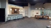 The Warehouse amenity space offers a thoughtfully designed, multi-zone environment ideal for entertainment and socializing. The layout encompasses several distinct areas, each with its own character while maintaining a cohesive industrial-chic design language throughout.

The private screening room is a standout feature, finished with deep teal and navy walls, a dark ceiling with recessed lighting, and wall sconces that create an immersive cinematic atmosphere. Plush, dark upholstered armchairs with built-in cup holders are arranged in a tiered configuration facing a large projection screen, with floor speakers enhancing the audio experience.

Adjacent lounge areas feature warm, textured tile accent walls in rich earthy tones that contrast with lighter gray walls and dark ceilings. Comfortable neutral sofas and armchairs are arranged around glass-top coffee tables, with a wall-mounted flat-screen TV providing a focal point. Decorative lamps, framed artwork, and teal glass accents add curated detail throughout.

The bar and game area includes a white-topped bar counter with industrial-style pendant lighting, bar stools, and a built-in media niche with tile surround. A billiards table anchors the game zone nearby. Carpeted flooring in a textured gray runs throughout, tying all zones together seamlessly.