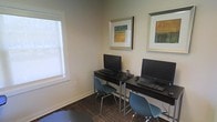 This single-floor space is designed as a functional business center, offering a focused and efficient environment for work and productivity. The room features two dedicated computer workstations, each equipped with a desktop computer, keyboard, and mouse, set on sleek dark desks paired with contemporary teal chairs. Natural light filters in through a frosted window, lending a bright yet comfortable atmosphere to the workspace. The neutral-toned walls provide a clean, professional backdrop, while two framed art pieces — featuring warm earth-toned abstract designs — add a welcoming aesthetic touch. The patterned carpet anchors the space and contributes to a quiet, concentrated work setting. The overall layout is thoughtfully organized to accommodate multiple users simultaneously while maintaining a sense of order and ease. Whether used for catching up on emails, printing documents, or general computing tasks, this business center is well-appointed to meet a variety of professional needs in a tidy, accessible setting.