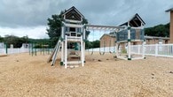 This outdoor playground offers a thoughtfully designed recreational space centered around a substantial wooden play structure finished in white and slate blue with green accent hardware. The centerpiece is a elevated clubhouse tower featuring a decorative peaked roof with a sunburst detail, providing an enclosed upper platform accessible by ladder. Extending from the main tower is a horizontal beam supporting multiple belt swings, connecting to a secondary elevated platform with railing on the opposite end. A straight slide descends from the main structure, while additional freestanding play equipment, including gymnastics-style rings and parallel bars, is visible along the perimeter. The entire play area is surfaced with wood chip mulch, providing a safety cushion underfoot. A white vinyl privacy fence borders the space, and mature trees provide a natural backdrop. The surrounding area includes a nearby residential building, indicating this playground serves as a community amenity. The overall design balances structural variety with safety-conscious materials, offering multiple play opportunities for a range of ages in an open, accessible outdoor setting.