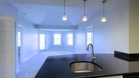 This single-floor residence showcases an open-concept layout that seamlessly connects the kitchen and living areas. The kitchen is anchored by a dark granite island countertop with an undermount sink and a sleek gooseneck faucet, offering a striking contrast against the light-toned surroundings. Three elegant pendant lights hang overhead, providing focused illumination over the island workspace.

The living area beyond features light carpeting and smooth, light-gray walls that contribute to a clean, airy atmosphere. A distinctive curved wall arrangement hosts multiple large windows, allowing abundant natural light to fill the space from several angles. This bay-style window configuration creates a welcoming, open ambiance while offering views to the exterior. An additional doorway with a side window is visible along the far wall, further enhancing the brightness of the room.

The transition between the kitchen and living space is fluid, making the layout well-suited for both everyday living and entertaining. The overall design balances functional kitchen features with a comfortable, light-filled living environment, unified by a consistent neutral color palette throughout.